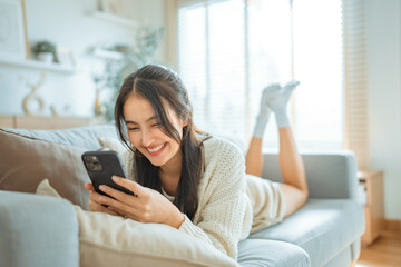 Happy young asian woman lady browsing surfing wireless internet on mobile phone while sitting a sofa couch in living room at home apartment, Shopping online via website