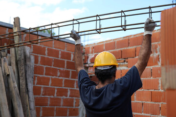Construction worker carrying an iron beam at a construction site. Builder working on construction site. Professional bricklayer working on house construction. Concept of architecture, industry.   - obrazy, fototapety, plakaty