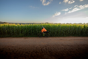 Ciclista de roupa laranja,  passando por Estrada rural, Plantação de Milho e céu azul