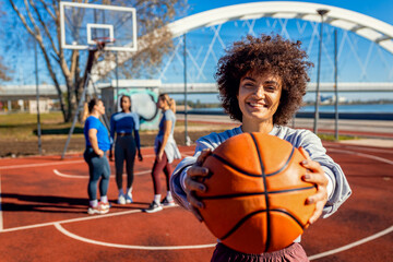 Portrait of young woman with her female friends on basketball court looking at camera and holding ball in her hands.