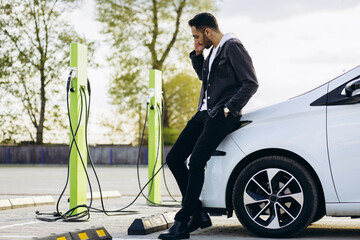 Man at charging station charging electric car and using mobile phone