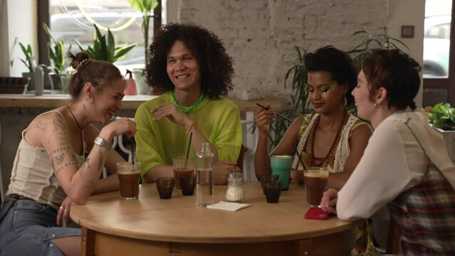 Lgbt people talk and drink alcohol at table in cafe. Young lesbian women, gay talking and looking with smiles, drinking cocktails and sitting in interior. Group of friends celebrate with emotion