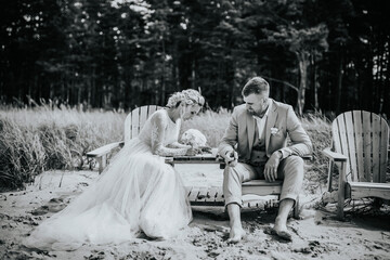Valmiera, Latvia - August 10, 2023 - Wedding couple seated on beach chairs in sand, looking at a phone, with forest in background.
