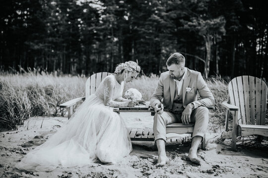 Valmiera, Latvia - August 10, 2023 - Wedding couple seated on beach chairs in sand, looking at a phone, with forest in background.