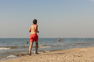 Man running topless barefoot on a sandy beach near the sea shore; male jogging on a sandy beach on a hot summer day during a heatwave.