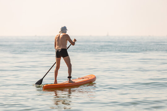 Senior man on a sup board on a calm sea; retired man standing on a paddle board in the sea on a summer day. - Powered by Adobe