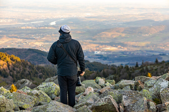 Photographer holding camera with long lens stands on rocky mountain and looks out over river valley with hills, settlements and forest; man with camera admires beautiful view from top of rocky hill.