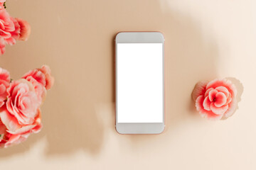 Smartphone with white screen and pink flowers with sunlight shadows on beige background, top view. Mobile phone mockup with copy space for design or web site promotion