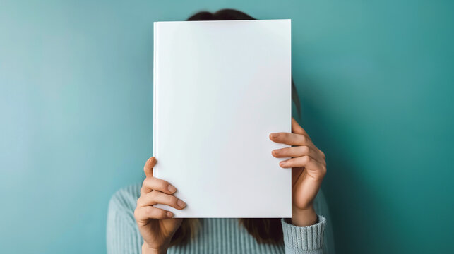 Woman holding blank white book cover in front of her face against teal background. Empty paper with copy space for text - Powered by Adobe