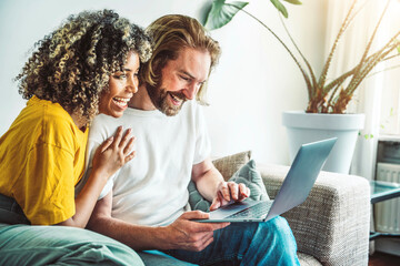 Multiracial young couple watching computer laptop sitting on the sofa at home - Happy diverse husband and wife using pc online services - Technology life style concept