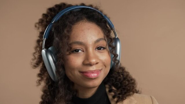 Positive mixed race woman listening music, enjoying headphones, beige background