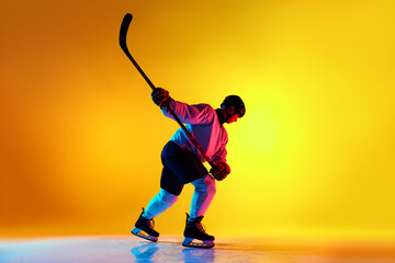 Naklejka premium Game-ready hockey player, geared up and focused, glides with stick held high in neon light against yellow gradient background. Concept of sport, competition, energy, tournament, healthy lifestyle. Ad