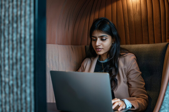 An Indian businesswoman engages in a virtual meeting using a laptop in a privacy booth, showcasing her adaptability and technological proficiency in professional settings.