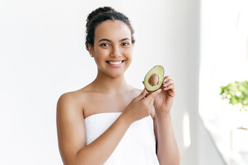 Beauty concept. Beautiful charming well-groomed brazilian or hispanic young woman wrapped in a towel stands on a white background, holds an avocado in her hands, looks and smiles at the camera