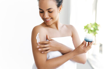Applying cream. Body skin care routine concept. Close-up photo of a hands of young hispanic or brazilian woman wrapped in a white towel, applying moisturizing cream on the shoulder, smiles