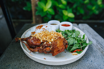 Deep fried duck with rice, green vegetables, and two types of sambals called Sambal Matah and Sambal Tomat on a white plate. Indonesian traditional cuisine. Food photography with bokeh background.
