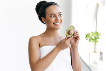 Beauty concept. Beautiful charming well-groomed curly haired brazilian or hispanic young woman wrapped in a towel stands on a white background, holds an avocado in her hands, looks away and smiles