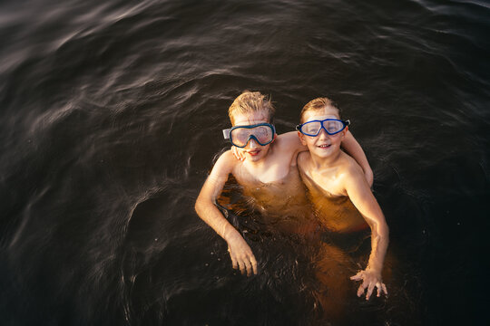 Two boys in swim masks looking at camera, hugging swimming into forest lake at sunset.