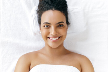 Top view of a beautiful well-groomed brazilian or hispanic young woman wrapped in a towel, lying in a beauty salon before a massage or facial skin care procedure, looking at the camera, smiling