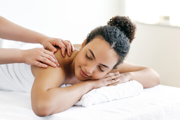 Relaxed satisfied brazilian or hispanic young woman lies et salon during a back and shoulder massage. Female massage therapist doing general body massage to a beautiful girl. Massage concept