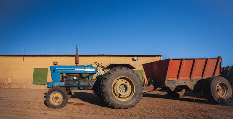 An old blue wheeled tractor with trailer stands in rural yard.