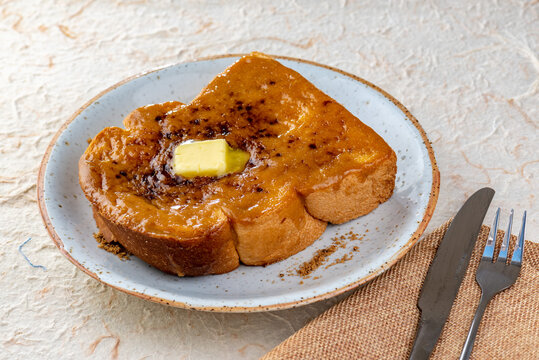 A photo of a single slice of French toast on a blue plate with butter on top and a fork and knife on the side.