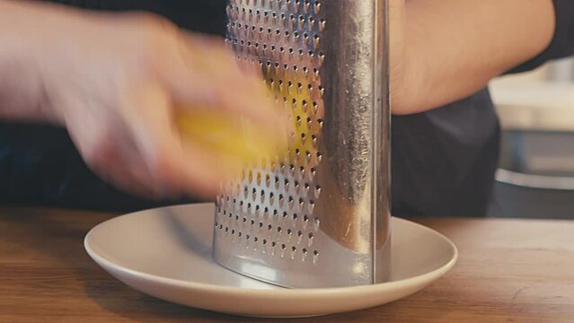 Grating fresh lemon peel, lemon skin with metal grater, on a white plate, on a wooden table in the kitchen. Interior. Studio lighting. Close up. High quality 4k footage