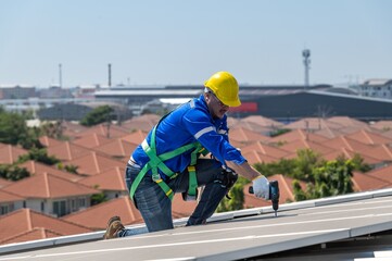 A solar panel technician in a yellow hard hat kneels to inspect a solar panel on a rooftop.