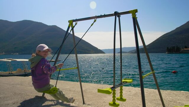 Close-up of a little girl riding on a swing on the seashore and admiring the beautiful view. Side view. Perast, Montenegro.