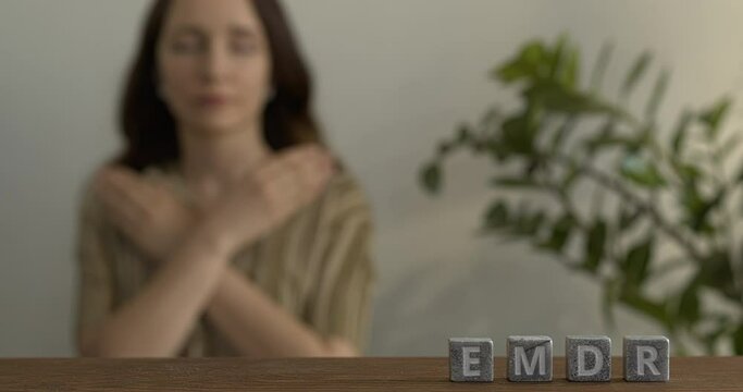 Letters EMDR written on grey stone cubes blocks. Female touching and tapping her shoulders in blurred background. Eye Movement Desensitization and Reprocessing psychotherapy treatment concept.