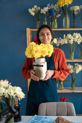 Florist woman holding a vase of daffodils while standing in her flower shop, looking at the camera and smiling. Small business and floristry concept