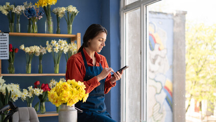 Hispanic female entrepreneur texting on smartphone while standing in flower shop. Happy employee typing on smartphone at work.