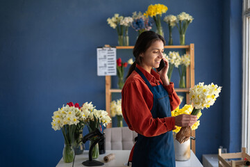 Side view of female florist in apron arranging fresh flowers for bouquet working in flower shop uses mobile phone for calling client