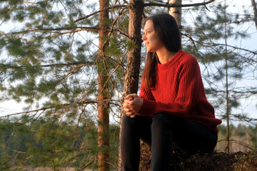 Smiling relaxed woman hiker sitting on hill admiring nature in mountain forest at sunset. Tourist traveler, camper, hiker enjoying hike outdoors. Calm female in red sweater in camping in woodland.