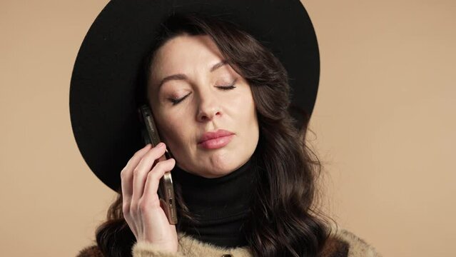 Pretty stylish woman talking by phone, smiling. Mature lady on beige background