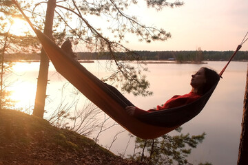 Calm relaxed woman lying in hammock in forest admiring lake nature at sunset. Resting relaxed young female in red sweater enjoying time in camping in dark woodland. Tourist, traveler, camper outdoors.
