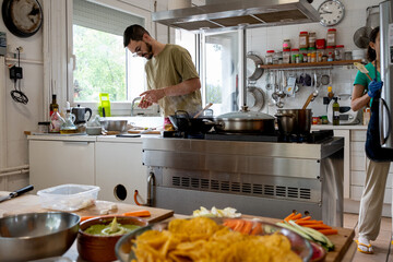 Young man cooking at the cooker, woman opening the fridge, in the foreground trays and dishes...