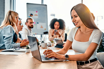 Brunette female using digital laptop on background of working space with diverse people. Attractive back end developer downloading new programs and connecting to server.