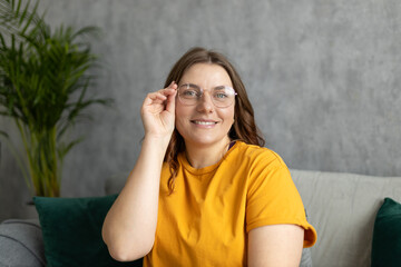 Close up face of young woman in glasses with beautiful smile siting on the sofa couch at home. 30s female rest and spend free spare time in living room indoors. People lounge concept
