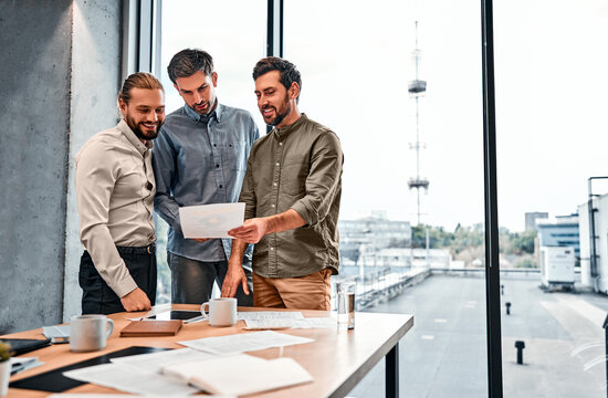 A team of different businessmen colleagues are standing in an office with panoramic windows, holding documents and discussing work issues. Copy space.