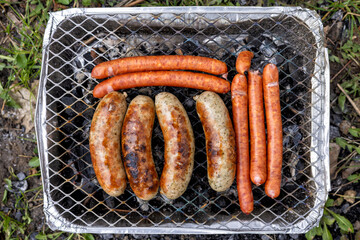 Sausages of various sizes are fried on the coals of a disposable grill.