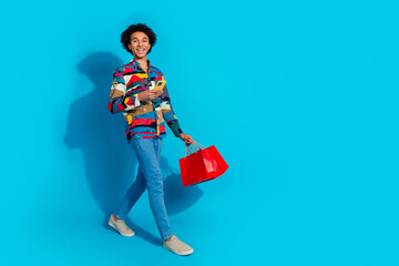 Full size photo of handsome young guy hold telephone walking shopping bags dressed stylish print garment isolated on blue color background