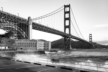 Panoramic wide angle view of iconic Golden Gate Bridge with view point ”Fort Point“ on a windy, sunny spring morning. High waves splash against the bank fortifications. Red monument seen from shore.