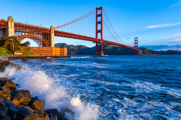 Panoramic wide angle view of iconic Golden Gate Bridge with view point ”Fort Point“ on a windy, sunny spring morning. High waves splash against the bank fortifications. Red monument seen from shore.