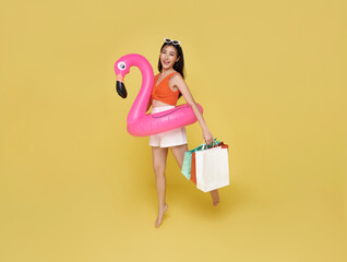 Happy sexy young woman dressed in swimwear holding shopping bag and flamingo rubber ring jumping for summer sale on yellow background.