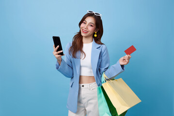 Beautiful Asian woman carrying shopping bags with credit card and mobile phone in hands on blue background.