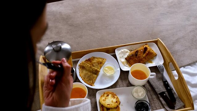 Woman is eating desserts in bed from wooden tray in hotel room on vacations, top view. Pancakes, cheesecakes, pastry with tea and ice-cream, condensed milk, sour cream and jam. Resting and relax.