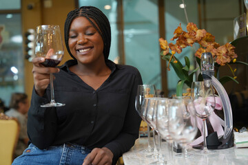Black female tourist sits holding a glass of red mattos wine, happily tasting wine at the counter bar in a wine shop. - obrazy, fototapety, plakaty