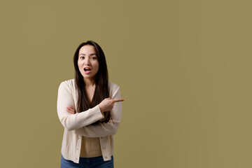 Shocked asian woman in casual clothes pointing with hand on background isolated on green 