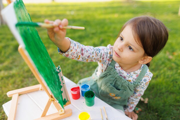 Little girl is painting a picture in the park on the green grass. Child development concept.